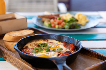 Side view of shakshuka with tomatoes, salami, chili pepper and fried eggs, served in hot cast pan