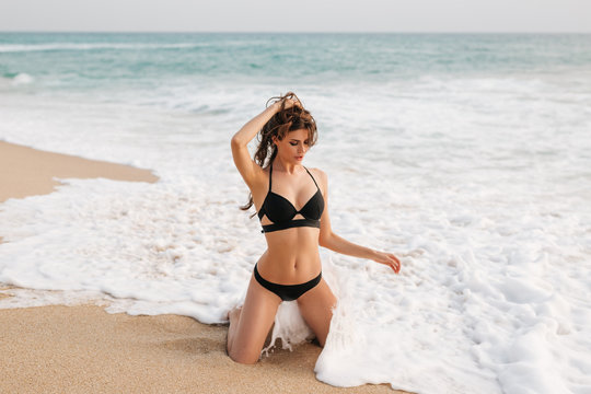 Woman in bikini swimsuit in sea waves on beach in summer. Girl model inblack swimwear with sexy body standing in water with wave splashing