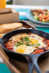 Close up of traditional israeli breakfast: shakshuka with tomatoes, salami, chili pepper and fried eggs, served in hot cast pan with toasts