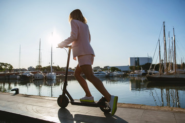Young beautiful woman in a pink suit riding an electric scooter in the port of the sea,  modern girl, new generation, electric transport, ecology, ecological transport, dawn, electric skateboard