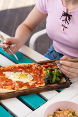 Woman is eating rome pizza with pepperoni, tomatoes and fried egg on a summer terrace