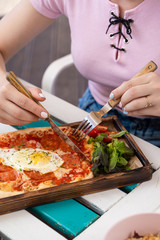 Woman is eating rome pizza with pepperoni, tomatoes and fried egg on a summer terrace