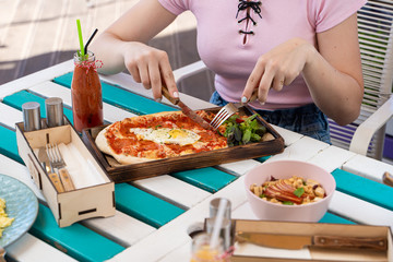 Woman is eating rome pizza with pepperoni, tomatoes and fried egg on a summer terrace
