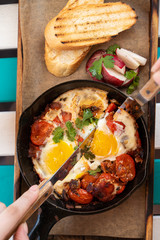 Woman is having breakfast on summer terrace: israeli shakshuka with tomatoes, salami, chili pepper and fried eggs, served in hot cast pan with toasts