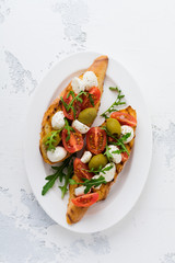Caprese bruschetta toasts with cherry tomatoes, mozzarella, olives and basil on white plate old light background. Top view.