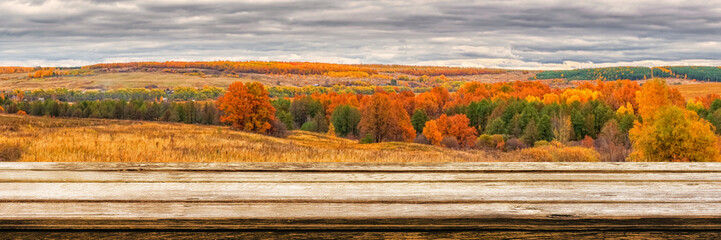 Empty wooden table with blurred picturesque autumn landscape of panoramic view from hill to lowland with field and grove in cloudy day. Mock up for display or montage products