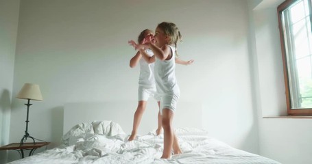 Authentic slow motion shot of two little girls sisters in white pajamas are having fun to jump on the bed of their parents bedroom. - Powered by Adobe