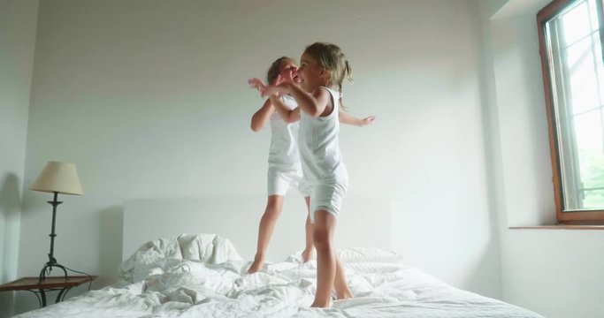 Authentic slow motion shot of two little girls sisters in white pajamas are having fun to jump on the bed of their parents bedroom. - Powered by Adobe