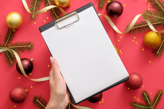 Hand holding clipboard mockup with blank paper over Christmas background with Christmas decorations, balls, gold ribbon, fir tree branches on red background. Checklist, New Year goals concept