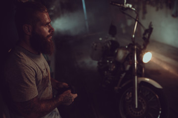 Fototapeta na wymiar Handsome brutal man with a beard is standing in his garage against the background of a motorcycle and looking away