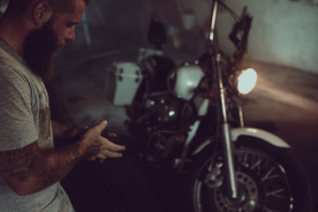 Fototapeta na wymiar Handsome brutal man with a beard is standing in his garage against the background of a motorcycle and looking away