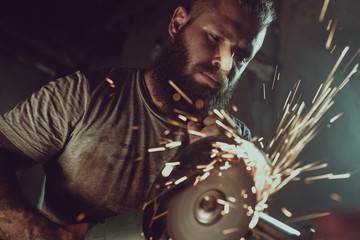 Handsome brutal male with a beard repairing a motorcycle in his garage working with a circular saw....
