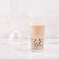 Tapioca pearl ball bubble milk tea, popular Taiwan drink, in drinking glass with straw on marble white table and wooden tray, close up, copy space.