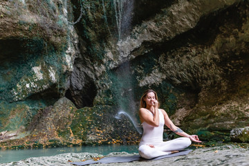 Calm and inspired young attractive slim fit girl sitting on a stone in the lotus position meditates against the backdrop of a mountain waterfall of green rocks and a river on a warm summer day