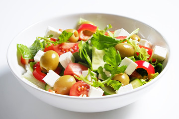 Traditional greek salad with fresh ingredients, feta cheese, olives, red tomatoes, cucumbers and greens in ceramic bowl, isolated on white background. Top view, close up, copy space.
