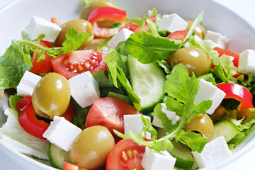 Traditional greek salad with fresh ingredients, feta cheese, olives, red tomatoes, cucumbers and greens in ceramic bowl, isolated on white background. Top view, close up, copy space.
