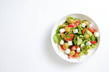 Traditional greek salad with fresh ingredients, feta cheese, olives, red tomatoes, cucumbers and greens in ceramic bowl, isolated on white background. Top view, close up, copy space.