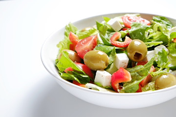 Traditional greek salad with fresh ingredients, feta cheese, olives, red tomatoes, cucumbers and greens in ceramic bowl, isolated on white background. Top view, close up, copy space.