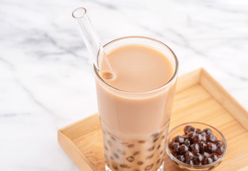 Tapioca pearl ball bubble milk tea, popular Taiwan drink, in drinking glass with straw on marble white table and wooden tray, close up, copy space.