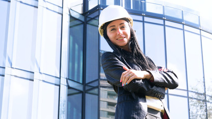 Business woman in suit and helment with crossed arms, looking at camera, smiling. Concept of: Skyscraper background, Communication, Manager, New business.