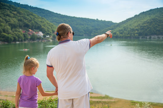 Father and daughter standing on the coast of the lake and holding hands. Father shows a hand forward to mountains. Bovan lake, Serbia. Travel, tourism concept.