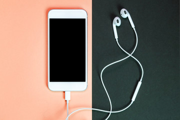 White smartphone on the orange and black background with headphones. View from above.