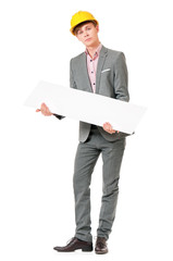 Full length portrait of young man in yellow safety helmet, holding empty board. Happy male with hard hat peeking from behind blank panel, isolated on white background. Guy showing blank placard board