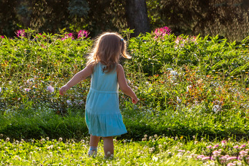 One beautiful little girl with long blonde hair in blue dress is in the summer garden among flowers.