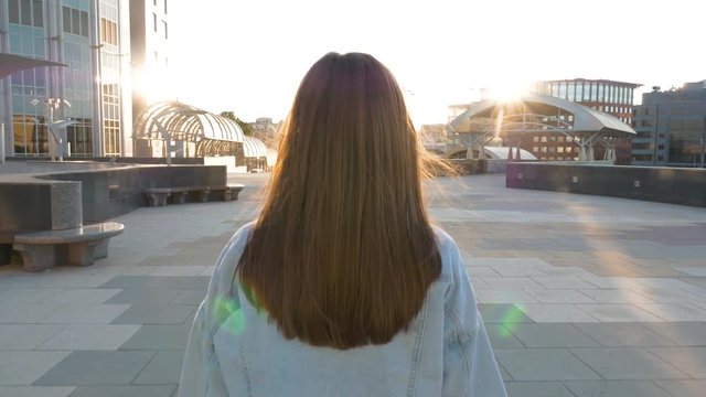 a young beautiful brown-haired woman with long hair walks towards the sun, along an empty street, during sunset. The camera follows her, view from the back. slow motion