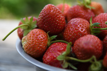  Strawberries in a bowl on a wooden table, outdoors on a green blurred background of leaves and trees
