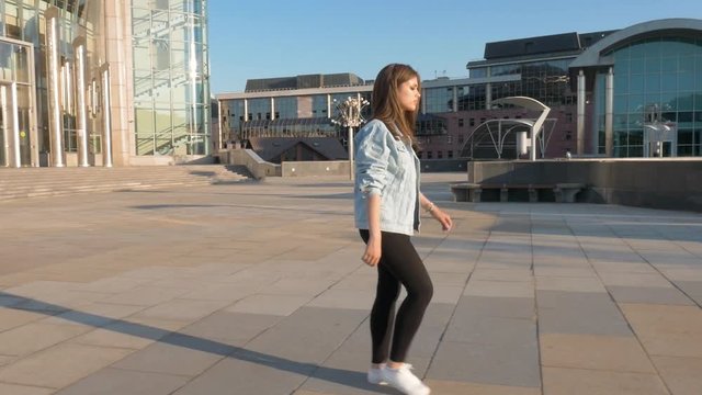 a young, pretty brown-haired woman in a blue denim jacket, black trousers and white shoes walks along the empty street towards the sun. the camera accompanies her. slow motion