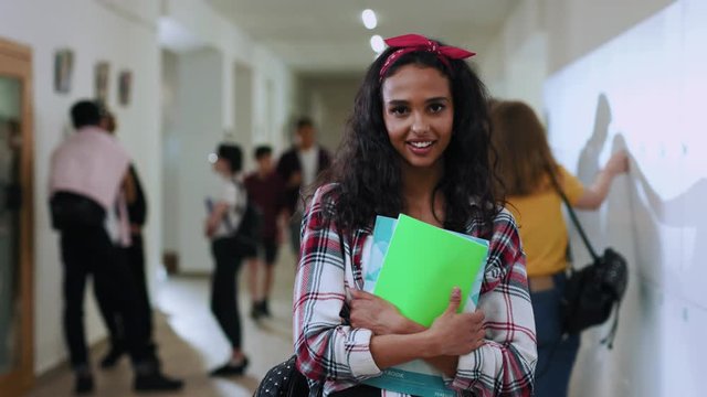 Amazing beauty latin teenage girl smiling on camera posing in school corridor during classes. Indoor portrait of cheerful schoolgirl with books standing by lockers in the corridor.