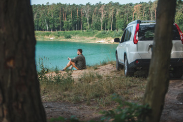 man sitting near white suv car at the edge looking at lake with blue water