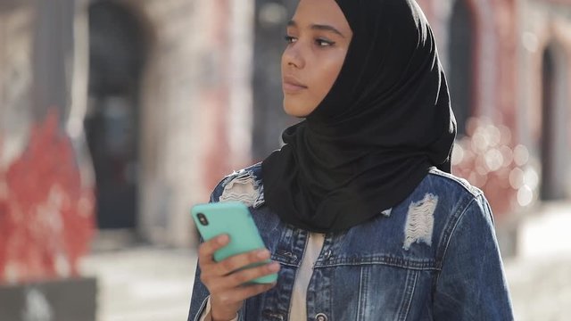 Young muslim woman wearing hijab headscarf standing in the city center and using smartphone. Communication, online shopping, social network concept.
