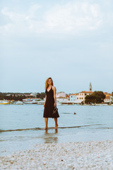 woman in black light dress walking barefoot by sea beach