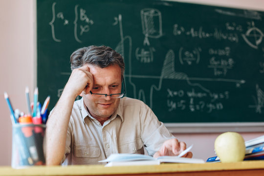 Middle age teacher in glasses attentively reading textbook in the classroom and smiling.- Image