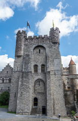 Fototapeta na wymiar Gent, Flanders, Belgium - June 21, 2019: Gray stone tower and adjacent buildings of Gravensteen, historic medieval castle of city against blue sky with white clouds. Flags on top, Green foliage.