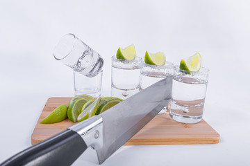 A knife and three shots of tequila with slices on top accompanied by slices of lemon and glasses on...