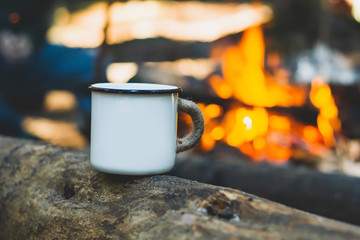 White enamel cup of hot beverage sitting on an old log by an outdoor campfire with a vintage folk edit. Selective focus on mug with blurred background.
