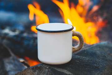 White enamel cup of hot beverage sitting on an old log by an outdoor campfire with a vintage folk...