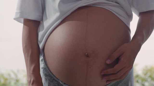 Young Asian Pregnant women show and looking ultrasound photo baby in belly. Mom feeling happy smiling peaceful while take care child lying near window in living room at home concept. Slow motion.
