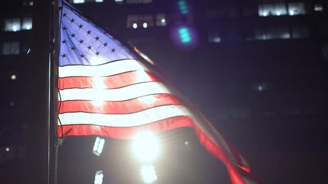 The United States flag in the wind at night in the city, backlit from bright lights.