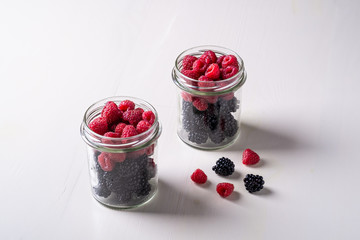 Raspberry and blackberry sweet organic juicy berries in two glass jars on white wooden table background
