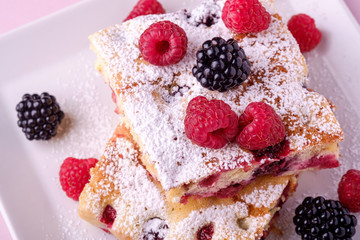 Homemade piece of cake, two pie slices with raspberry and blackberry berries, powdered sugar, in white square plate on pink paper background