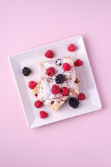 Homemade piece of cake, two pie slices with raspberry and blackberry berries, powdered sugar, in white square plate on pink paper background, top view, flat lay
