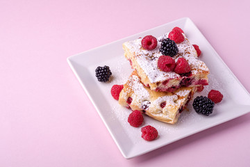 Homemade piece of cake, two pie slices with raspberry and blackberry berries, powdered sugar, in white square plate on pink paper background