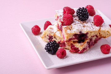 Homemade piece of cake, two pie slices with raspberry and blackberry berries, powdered sugar, in white square plate on pink paper background