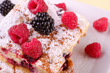 Homemade piece of cake, two pie slices with raspberry and blackberry berries, powdered sugar, in white square plate on cream yellow paper background, macro, close up
