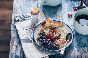 Healthy oatmeal breakfast with red currants, raspberry, grape and boiled egg with toast and coffee on a rustic blue background