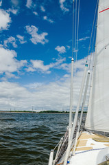 View of the water and the distant shore from the side of a sailboat with rigging and sails